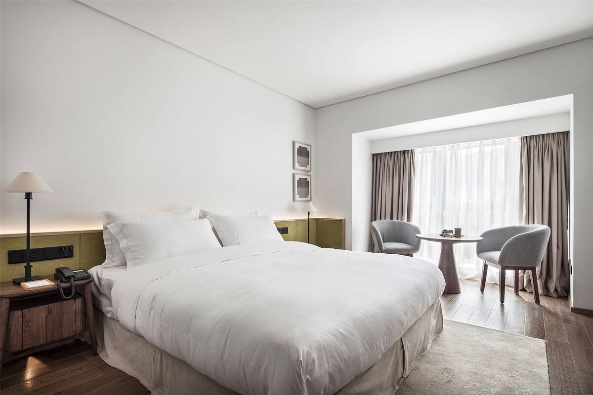 A hotel room with a large bed, bedside table with a lamp, a small table with chairs, and a window with curtains.