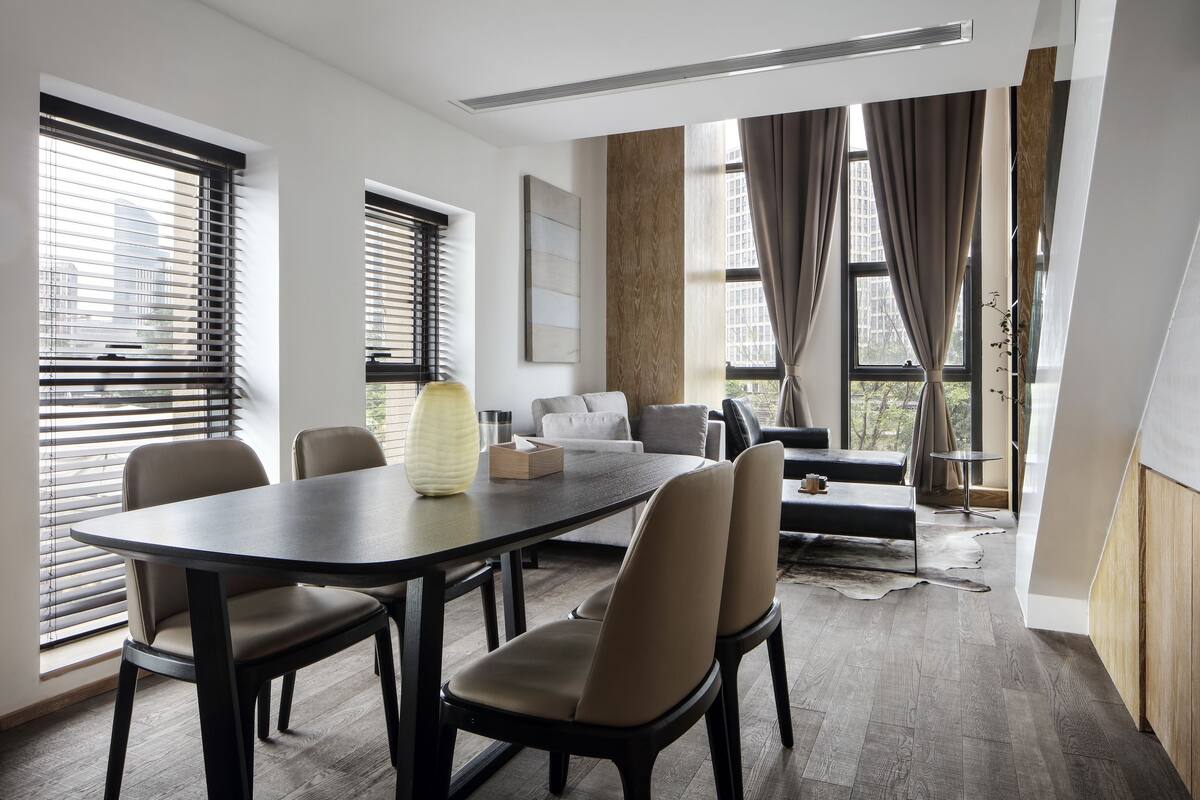 A modern dining area with a dark wooden table and chairs, large windows with blinds, and a view of the city.