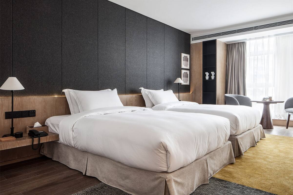 A hotel room with two beds, a bedside table with a lamp, a small table with a chair, and a large window with curtains.