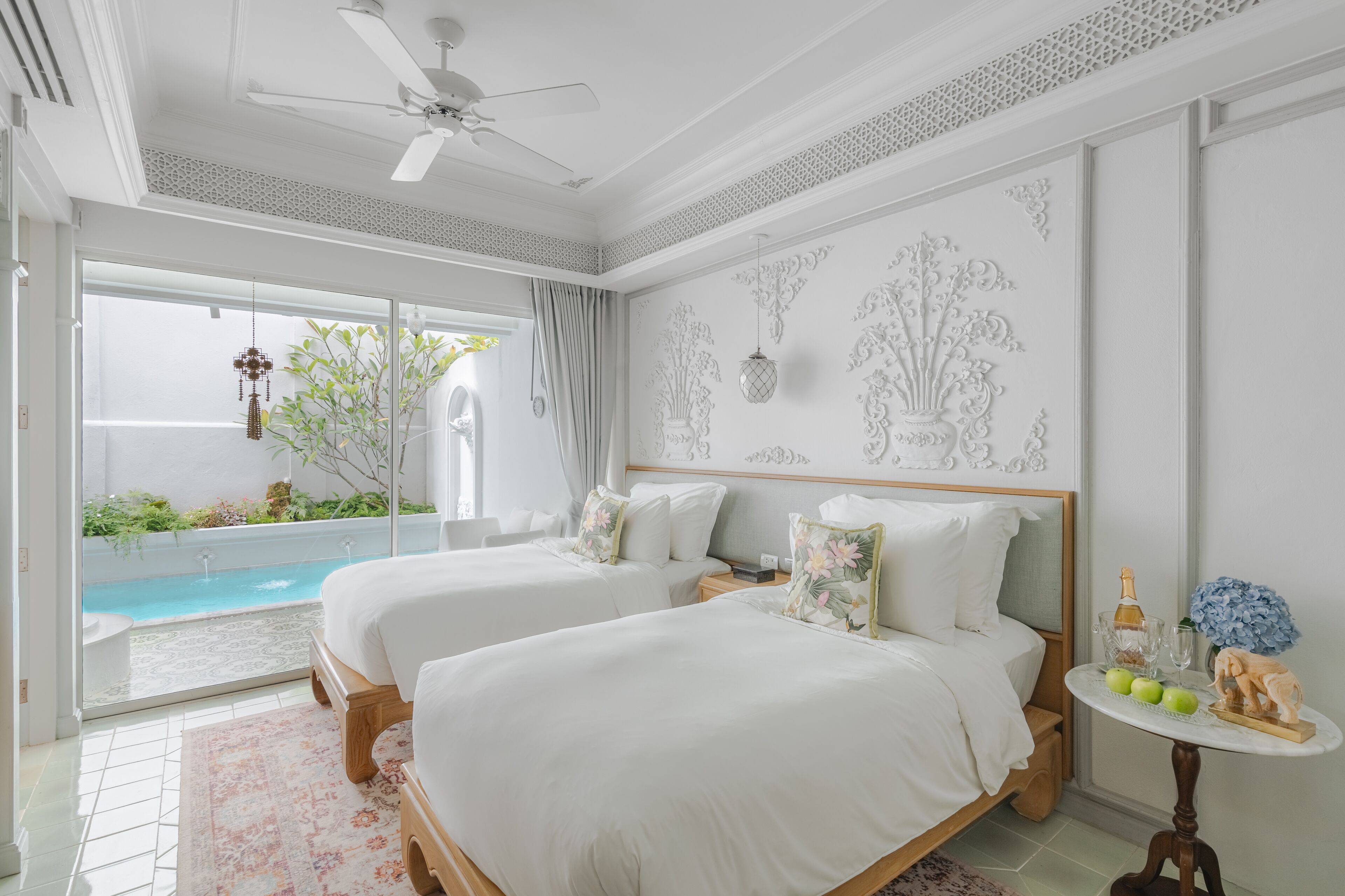 Habitación de hotel con dos camas, un ventilador de techo, una mesita con una botella y fruta, y vistas a una piscina.