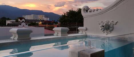 Una piscina en la azotea con agua azul cristalina, detalles arquitectónicos blancos y vistas al atardecer de fondo.