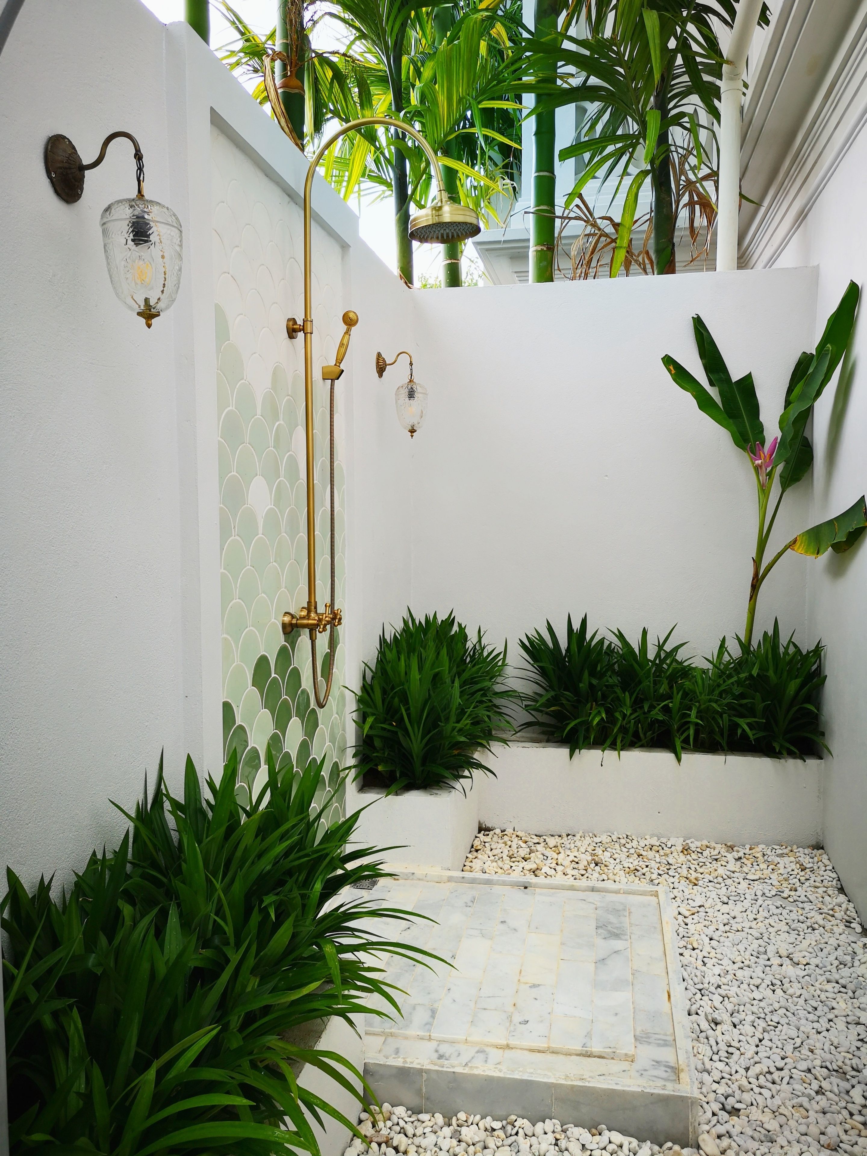 A small, enclosed shower area with a white tiled wall, a glass showerhead, and a gold shower control. There are green plants in white planters along the wall, and a small step leading to a pebble-covered ground.