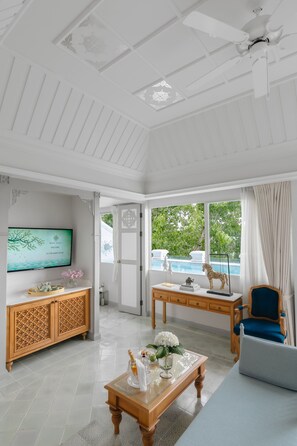 A spacious living room with a television, wooden furniture, a coffee table with flowers, and a view of a swimming pool through large windows.