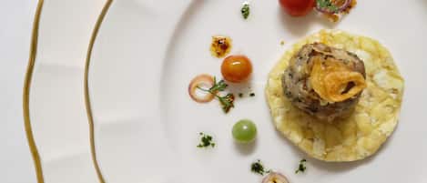Un plato blanco con borde dorado, que presenta un plato de carne con cobertura crujiente, adornado con hierbas y rodeado de tomates cherry pequeños y uvas verdes.