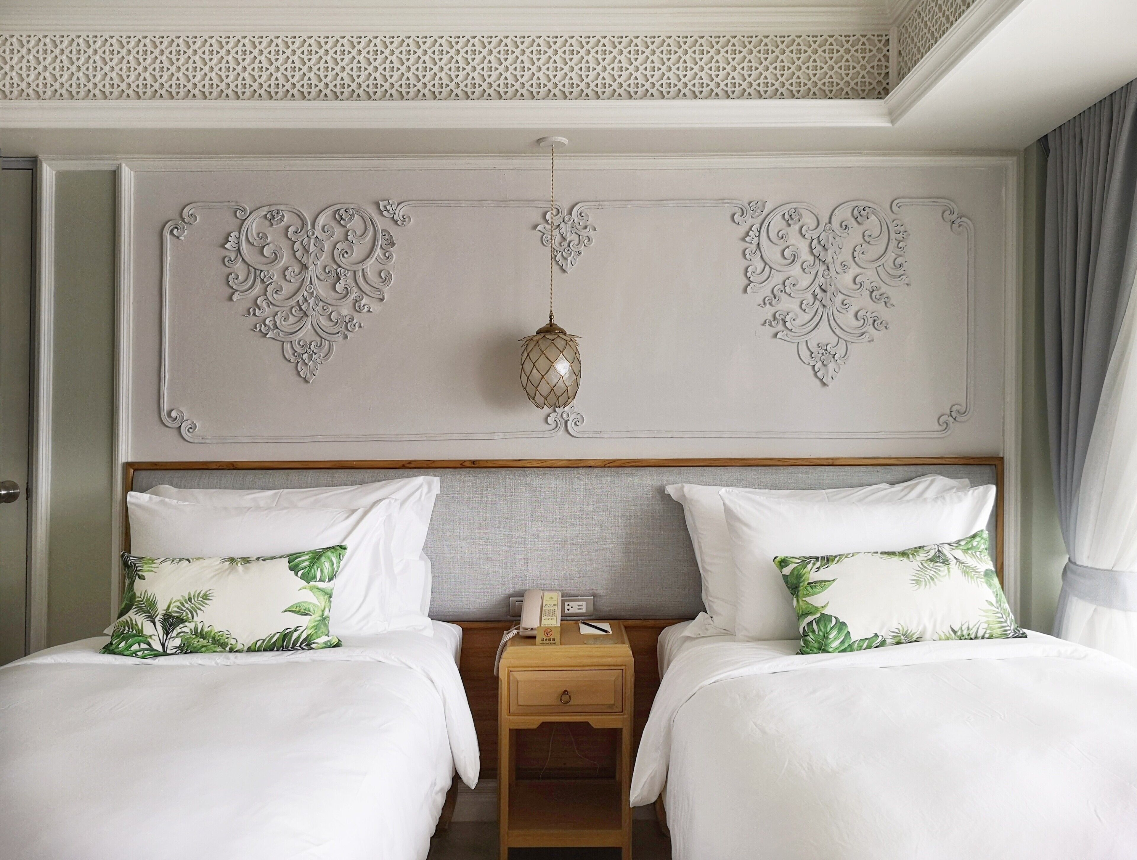 Een hotelkamer met twee bedden, een nachtkastje, een telefoon en een decoratief plafond met een hanglamp.
