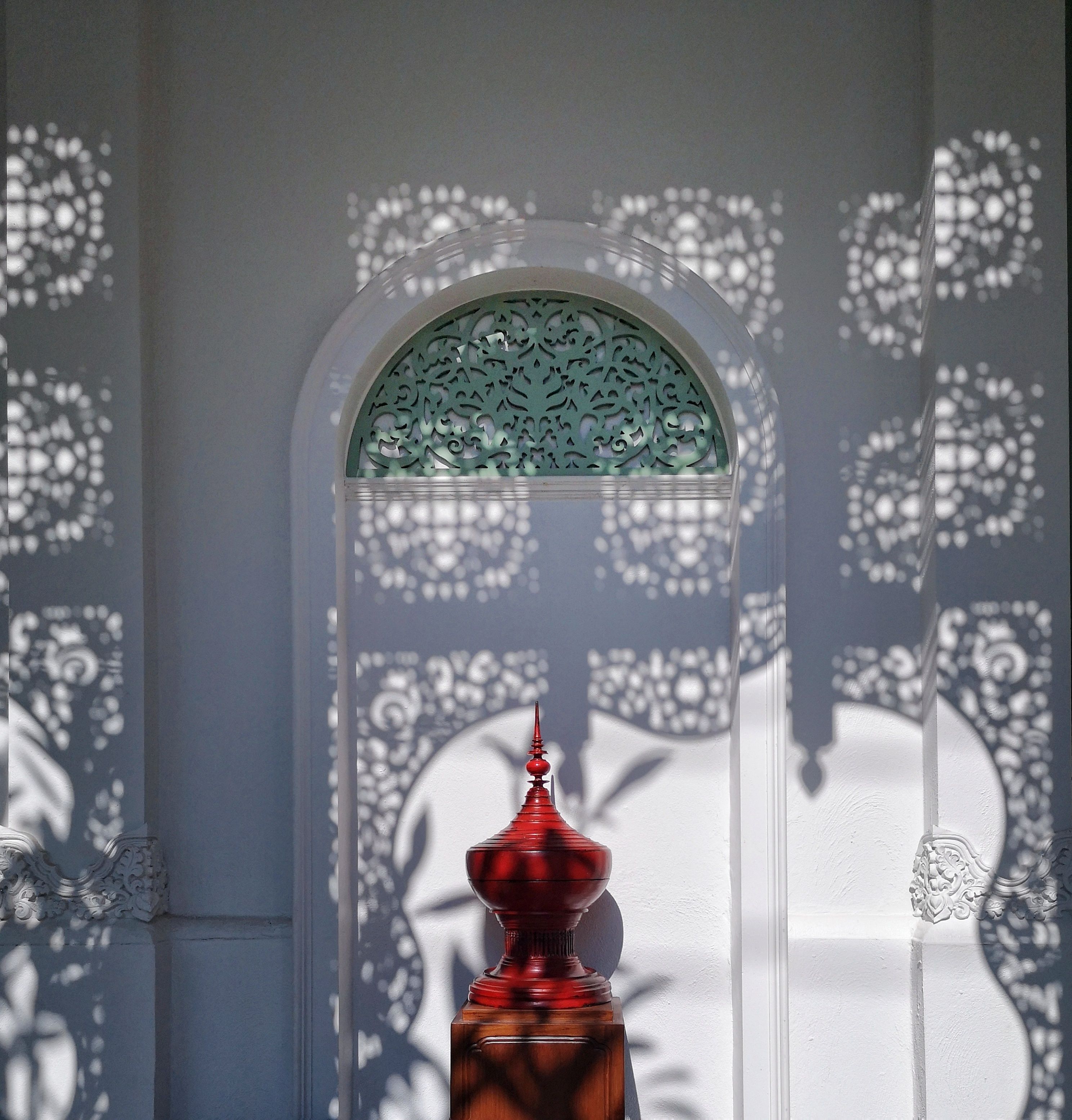 A room with a decorative window, a red object in the foreground, and intricate wall patterns.