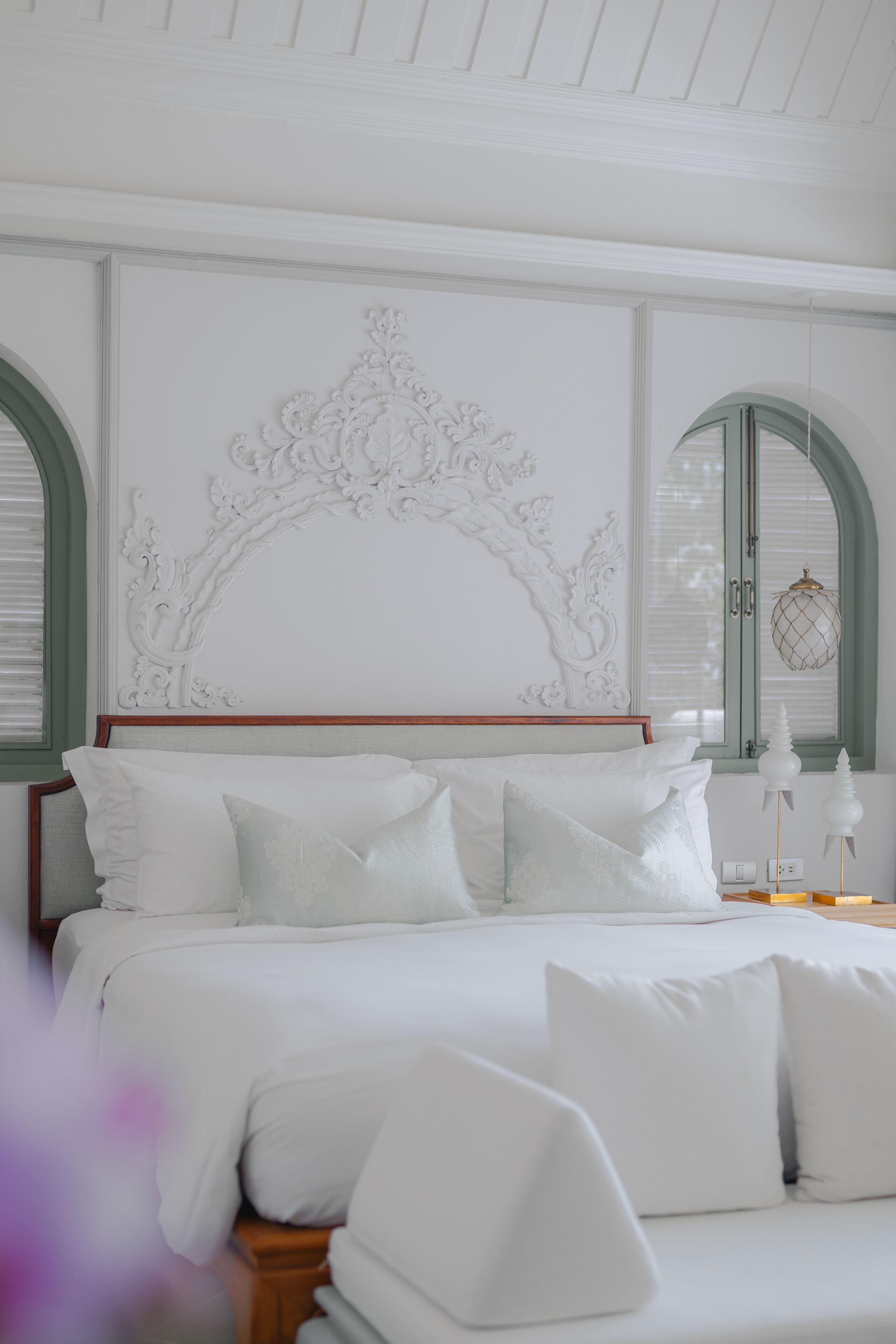 A neatly made bed with white linens and pillows, a decorative headboard, and two arched windows with blinds.