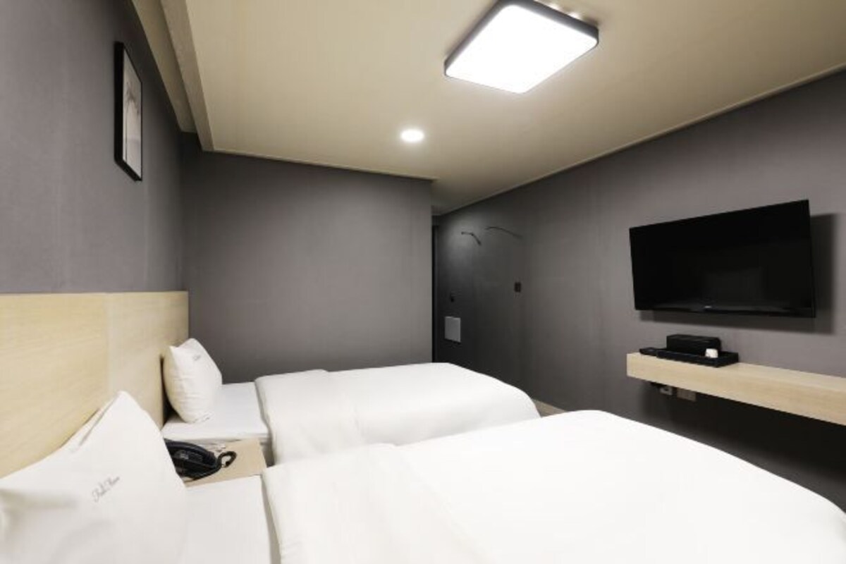 Habitación de hotel con dos camas, un televisor montado en la pared y una mesita de noche con lámpara.