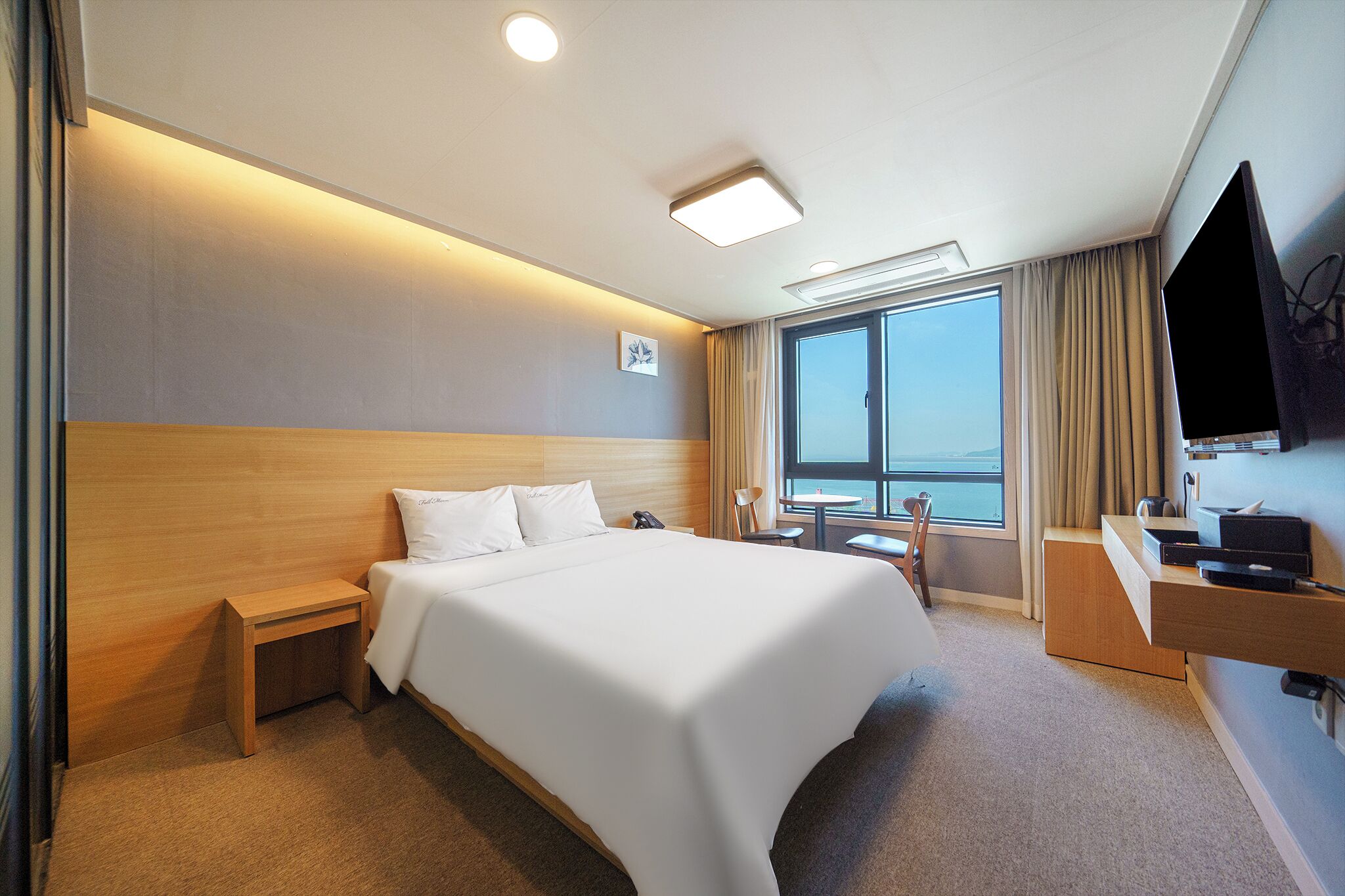 A hotel room with a large bed, bedside table, a desk with a chair, a TV, and a window with curtains.