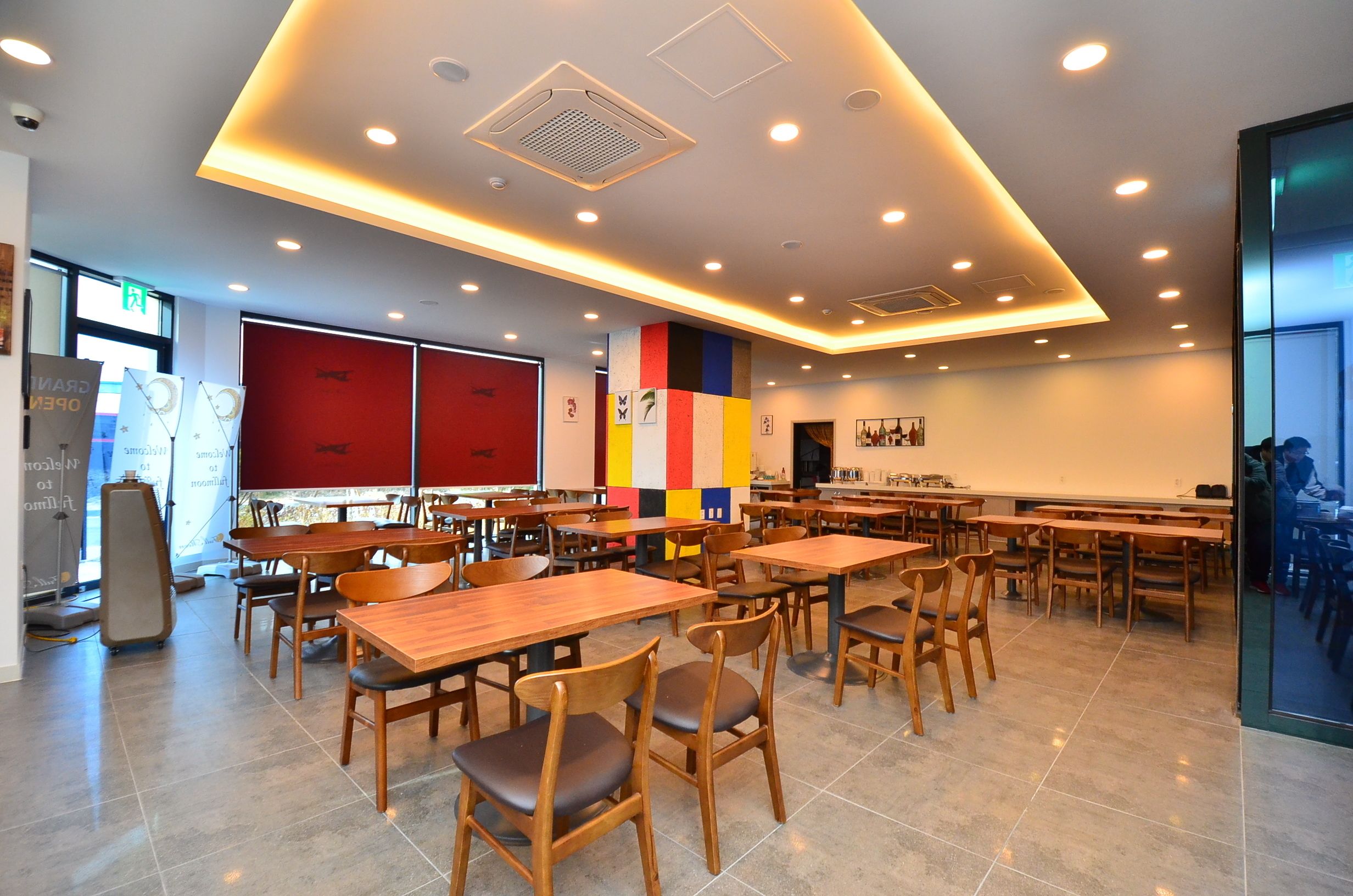 A modern dining area with wooden tables and chairs, a colorful wall, and recessed ceiling lighting.