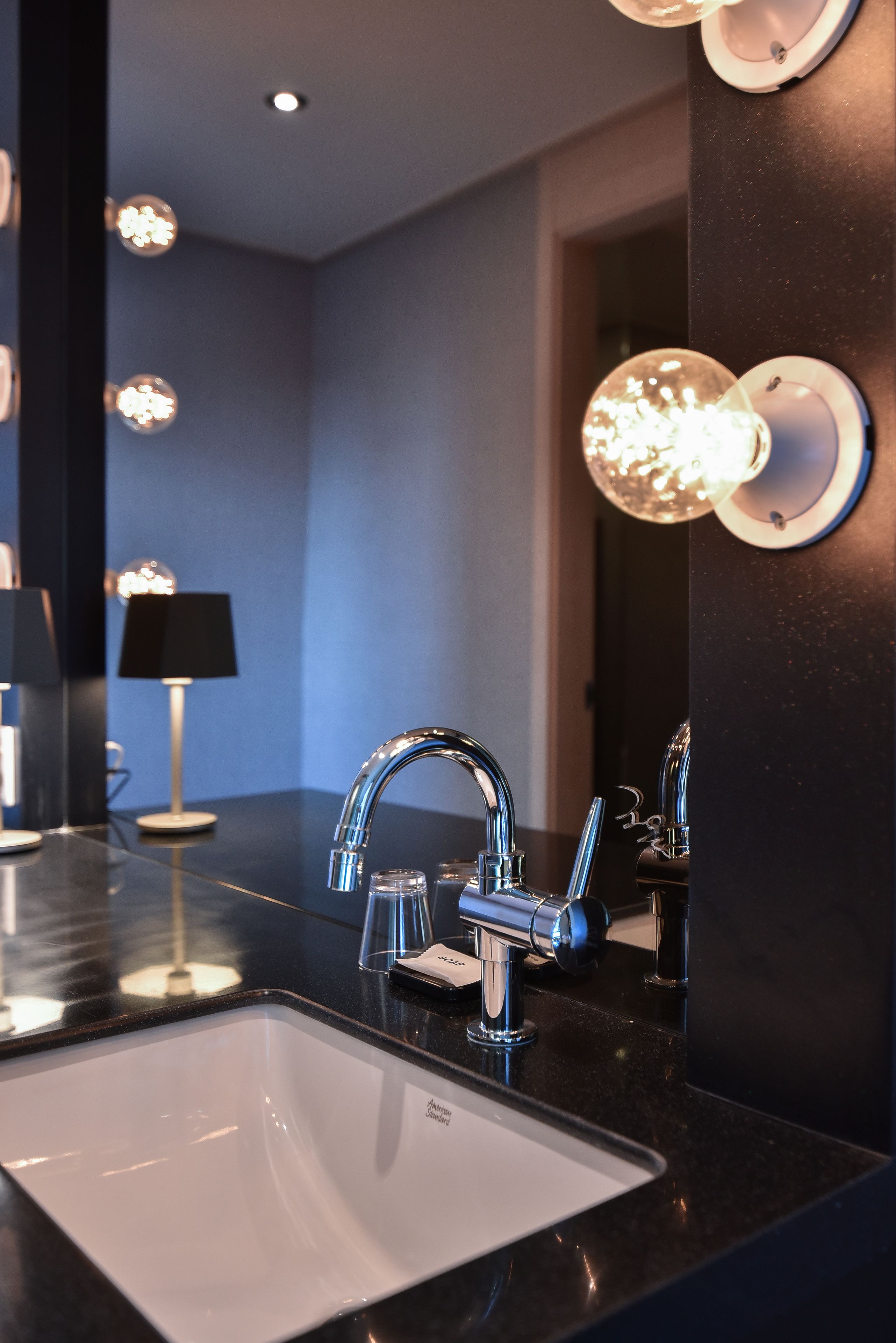 A modern bathroom with a black countertop, chrome faucet, and a white sink. There are wall-mounted lights and a mirror above the sink.