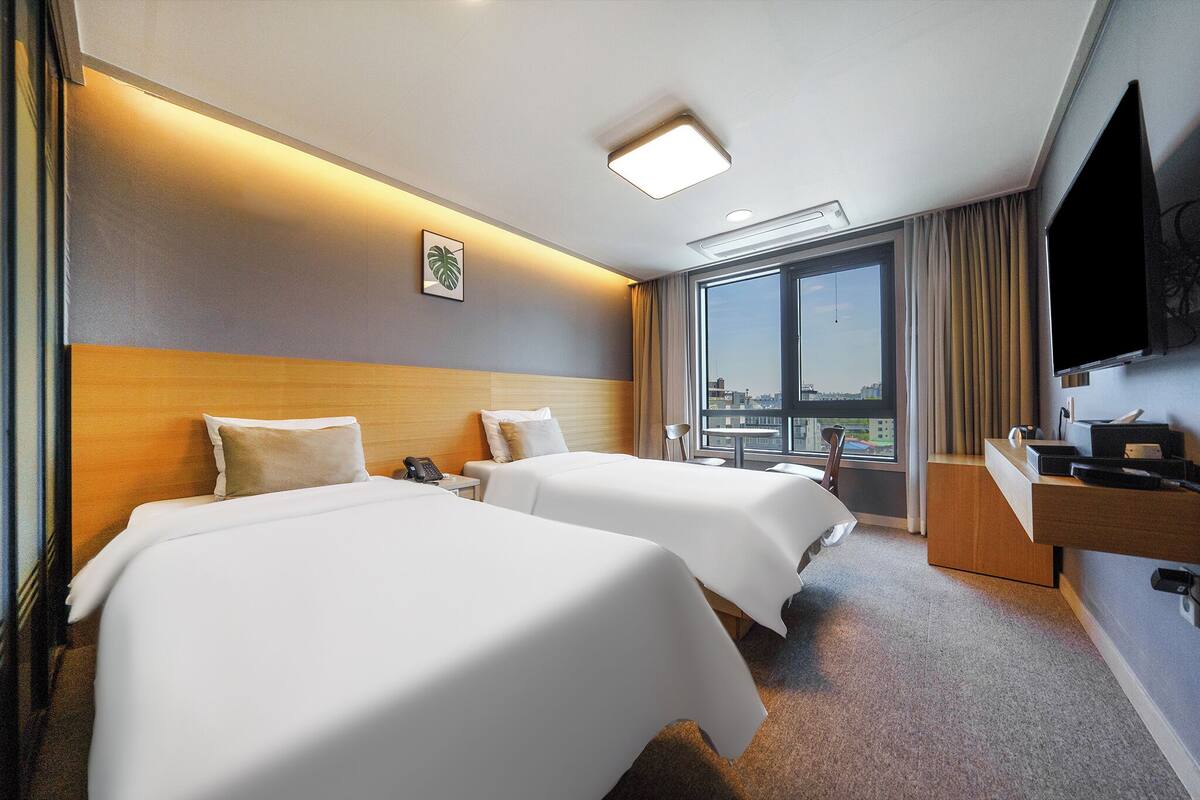 A hotel room with two beds, a TV, a desk, and a window with a city view.