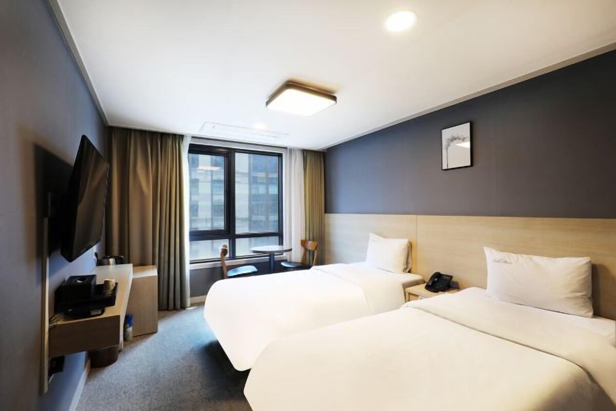 Habitación de hotel con dos camas, un televisor de pantalla plana, un escritorio con lámpara y una ventana con cortinas.