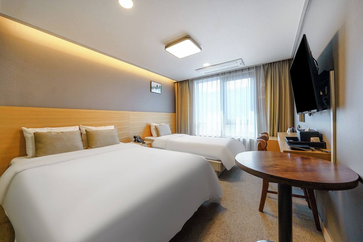 A hotel room with two beds, a desk, a TV, and a window with curtains.