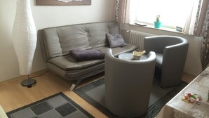 Un salon moderne avec un canapé gris, une table basse ronde et un petit fauteuil. Un tableau représentant un coucher de soleil est accroché au mur, ainsi qu’une lampe sur pied et une fenêtre avec des rideaux.