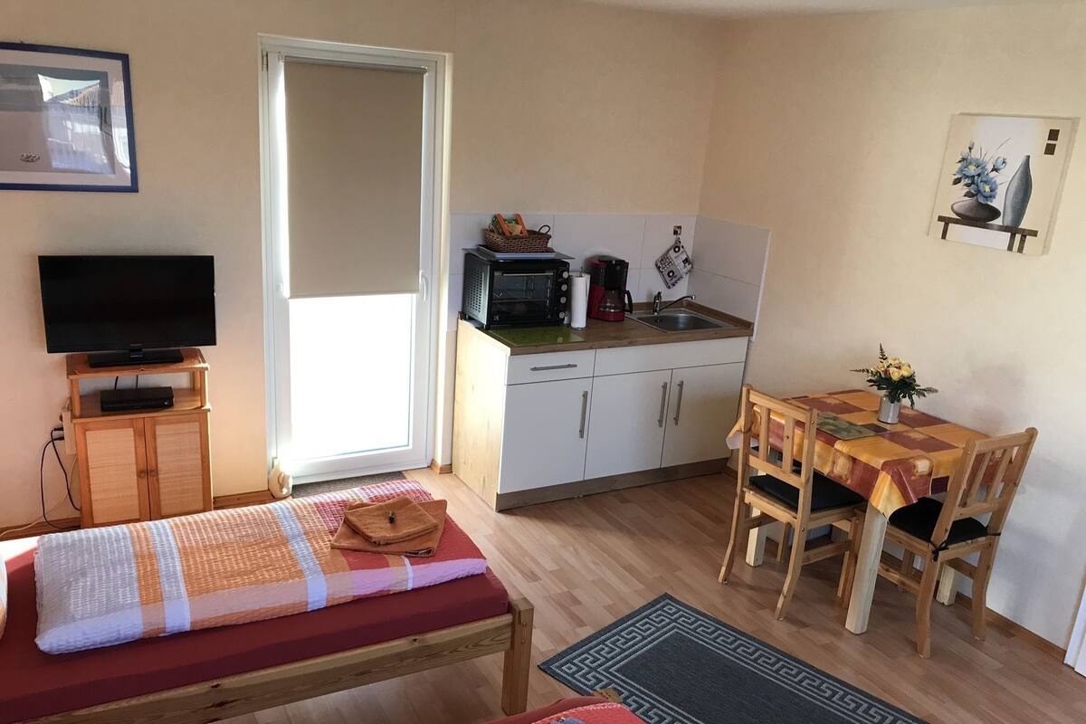 Un espace de vie compact comprenant un lit, une petite table avec des chaises, un coin cuisine et un téléviseur.