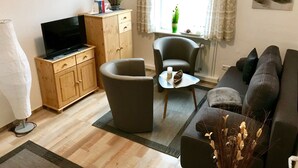 Un salon comprenant un canapé, un fauteuil, une table basse, un téléviseur posé sur un meuble en bois et une fenêtre avec des rideaux.