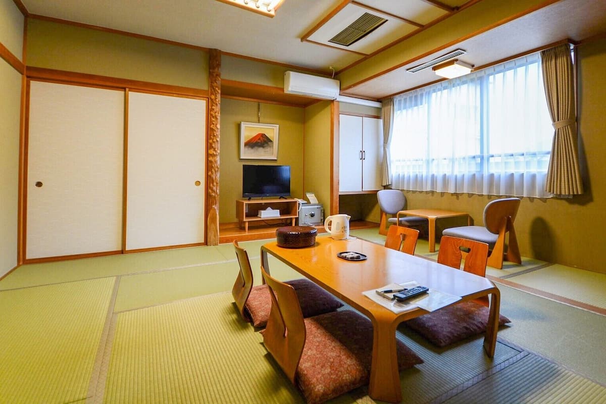 A traditional Japanese-style room with tatami flooring, a low table with cushions, a television, and a window with curtains.