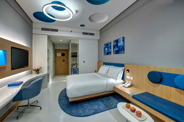 A modern hotel room with a bed, a desk, a TV, and a seating area.