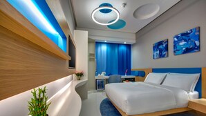 A modern hotel room with a curved white console, a bed with white linens, a small potted plant, a desk with a chair, and a blue accent wall with abstract art. - Al Khoory Inn (Dubai)