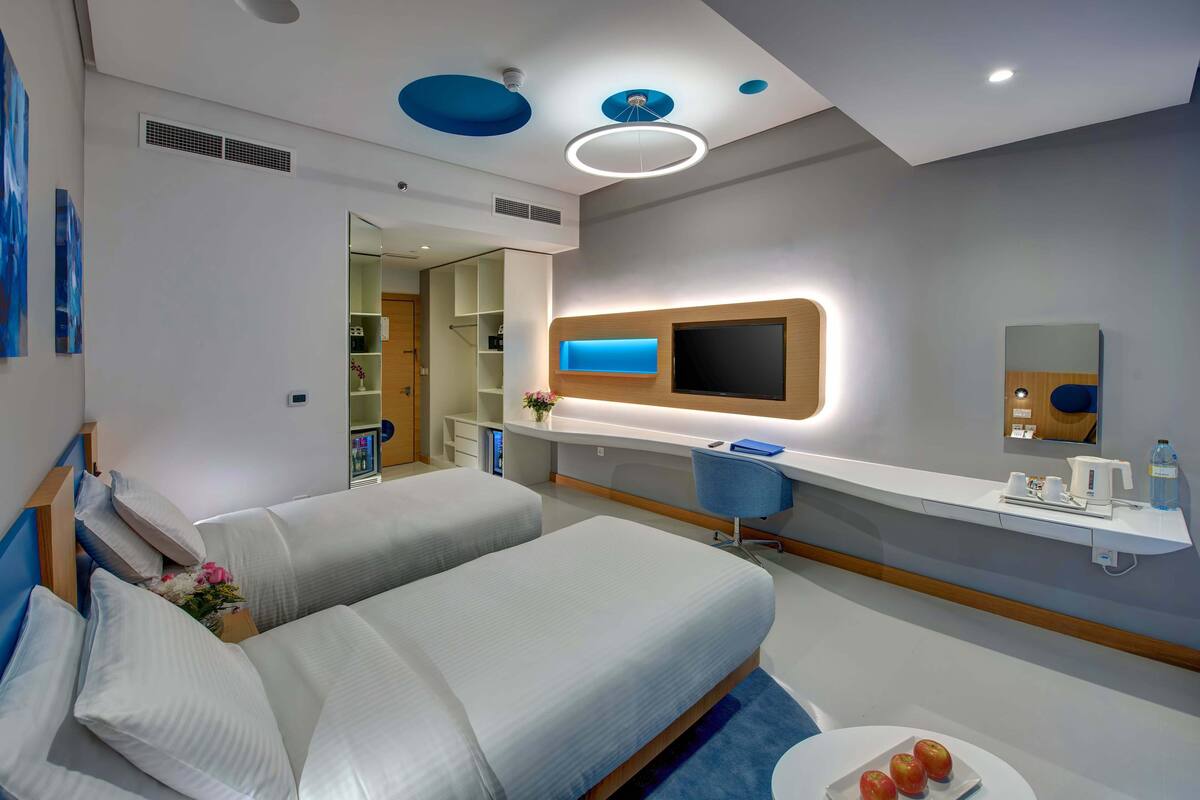 A modern hotel room with two beds, a desk, a TV, and a bathroom visible through an open door.