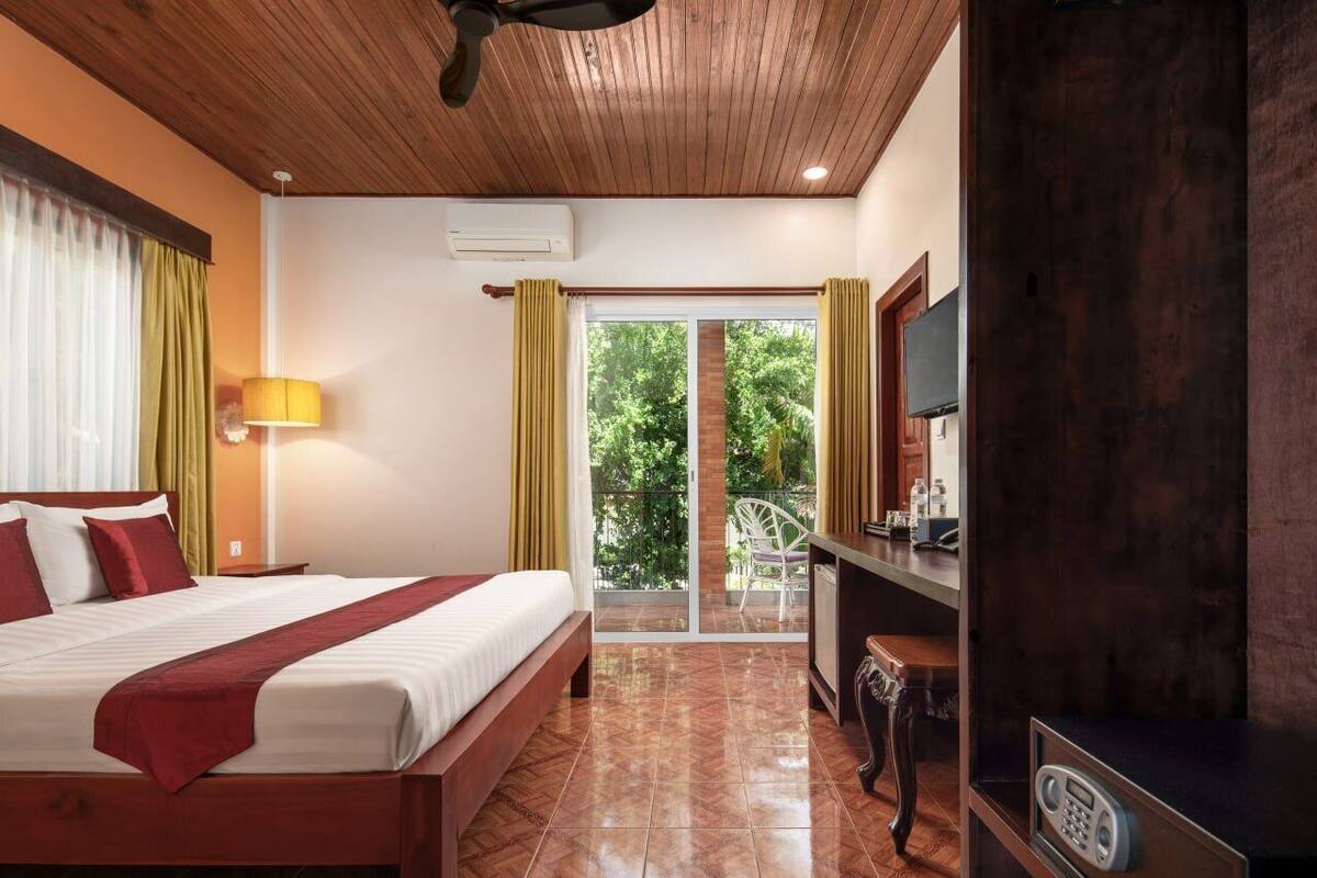 A hotel room with a wooden ceiling, a bed with red bedding, a desk with a chair, a TV, and a sliding glass door leading to a balcony.