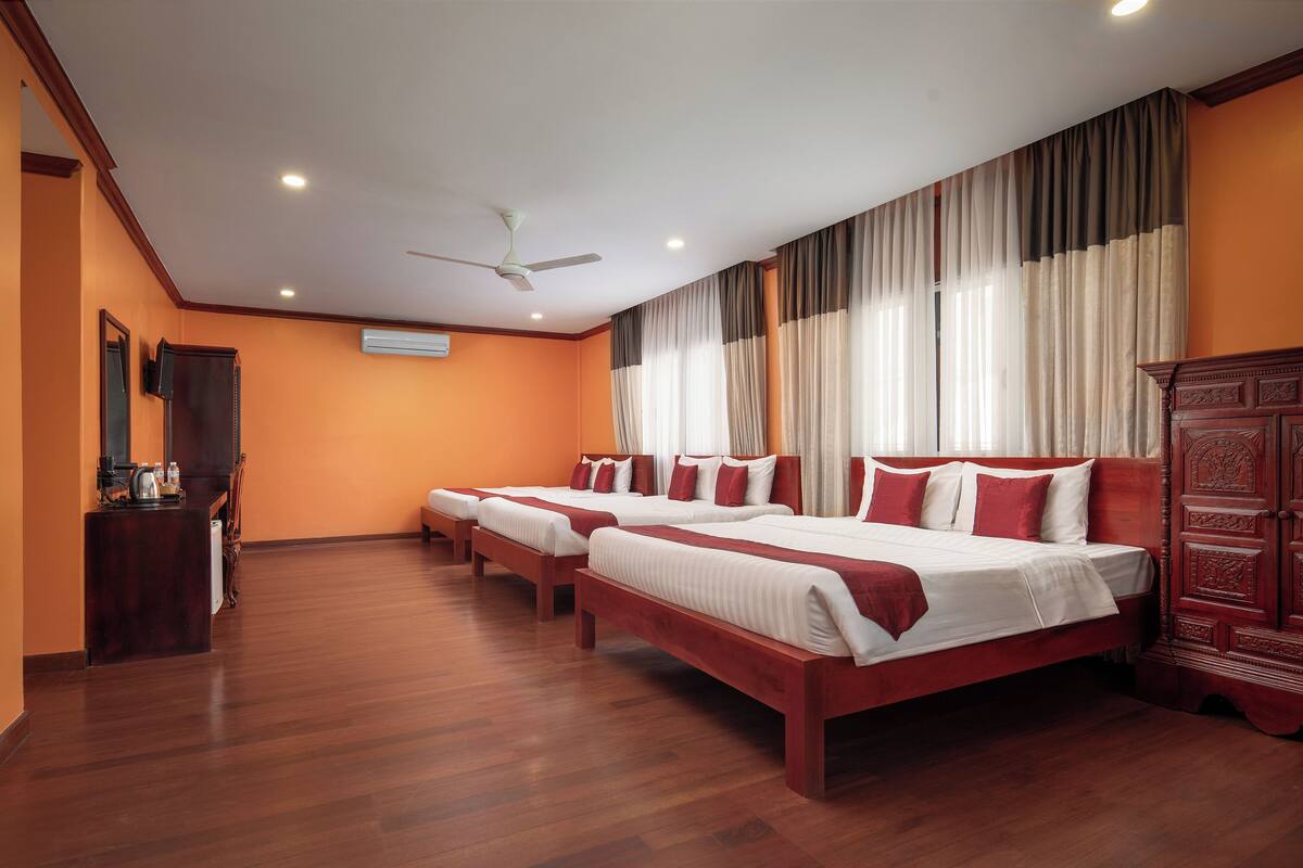 A hotel room with three beds, wooden flooring, a ceiling fan, and a large window with curtains.