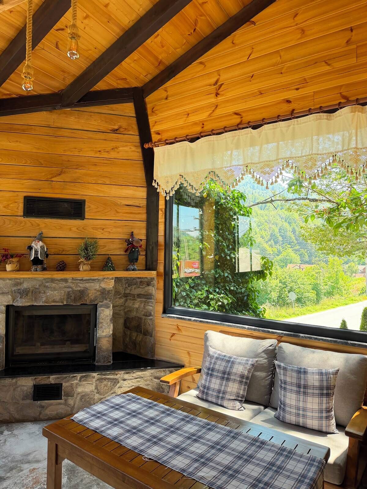 L’intérieur d’un chalet accueillant, avec une cheminée en pierre, des poutres en bois au plafond, un canapé agrémenté de coussins à carreaux et une vue sur la verdure environnante.