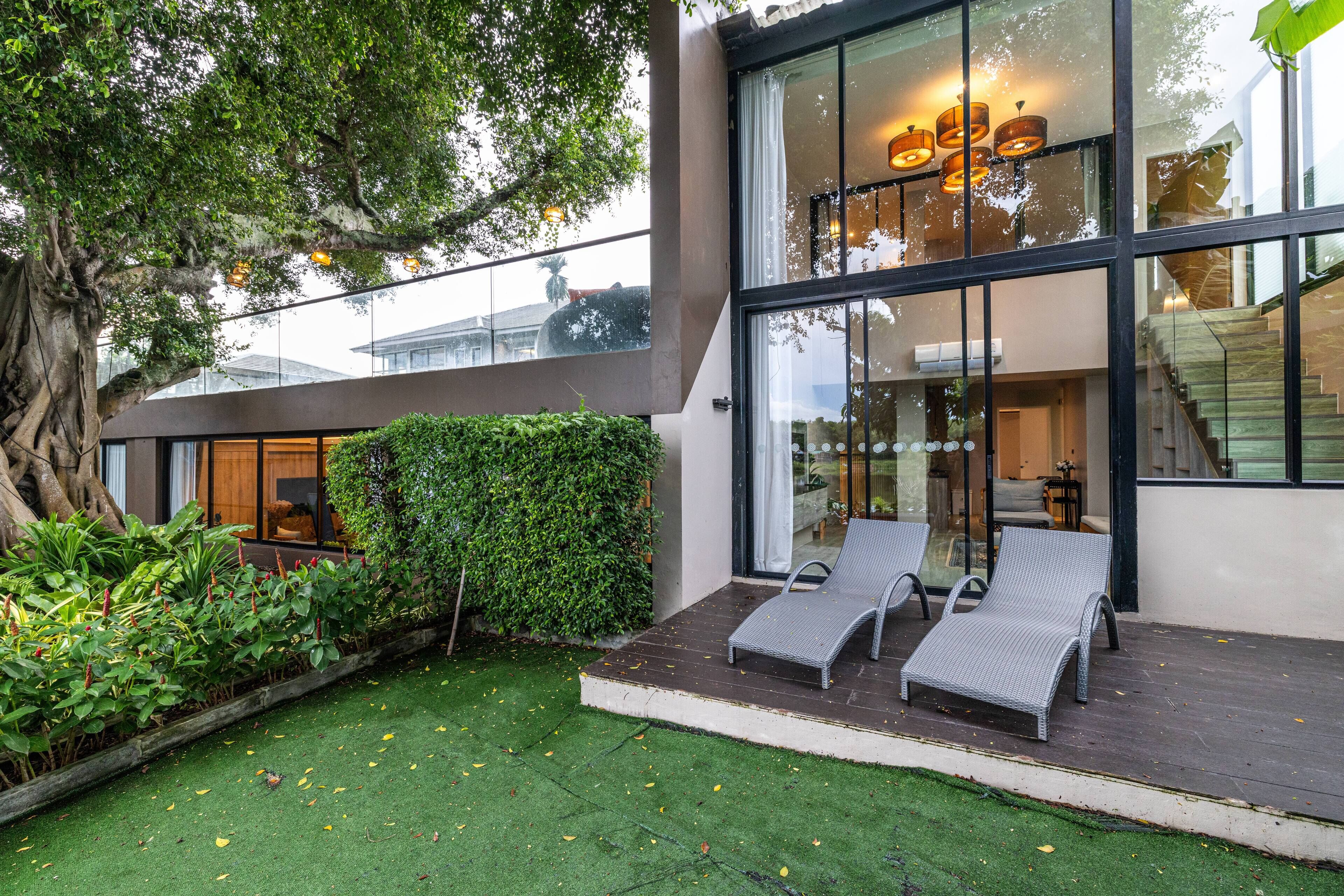 Uma casa moderna com fachada de vidro, um deck de madeira com duas poltronas de descanso e um jardim verdejante.