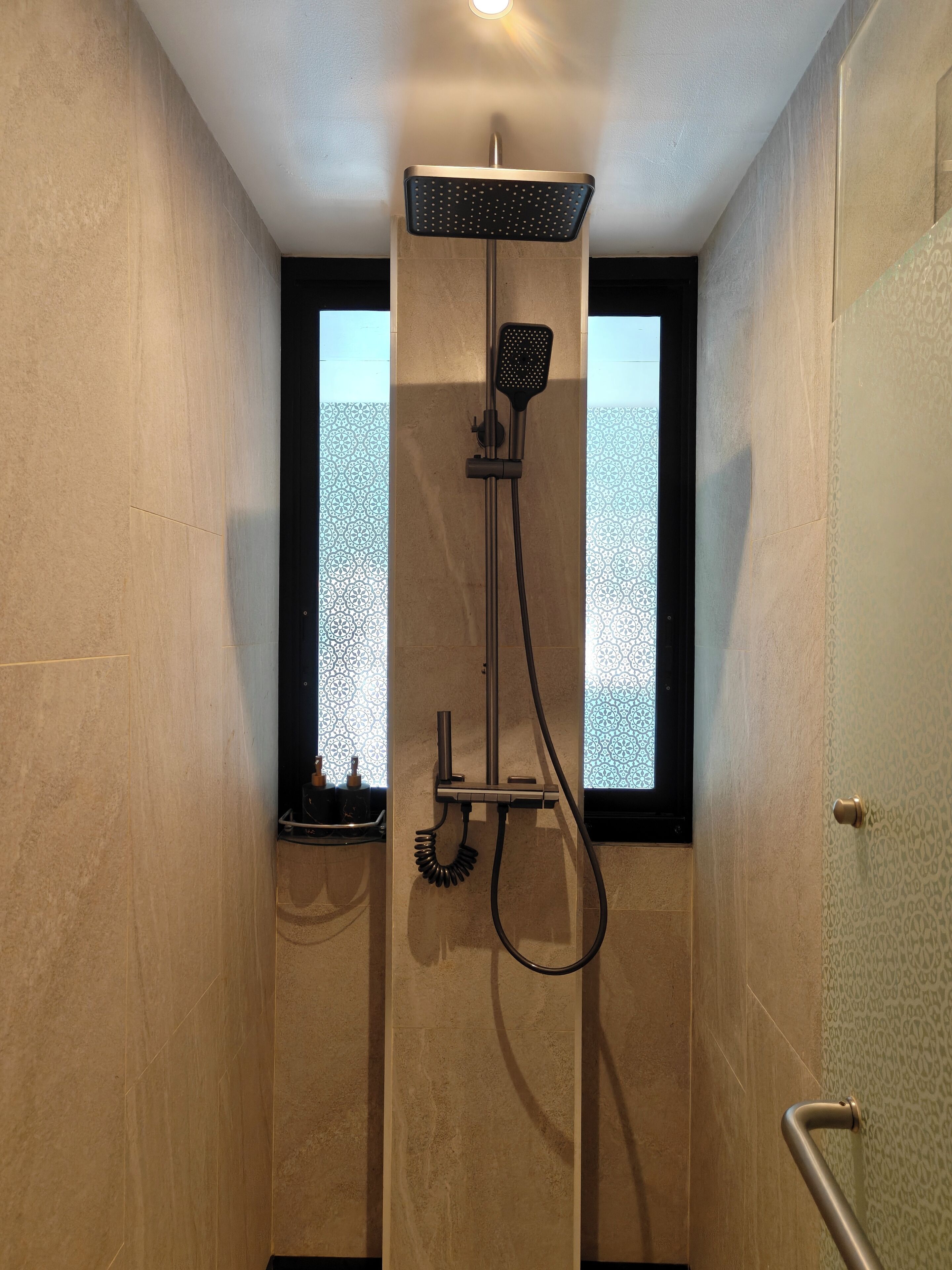A modern shower with a handheld showerhead and a fixed overhead showerhead, a window with a decorative pattern, and a towel rack.