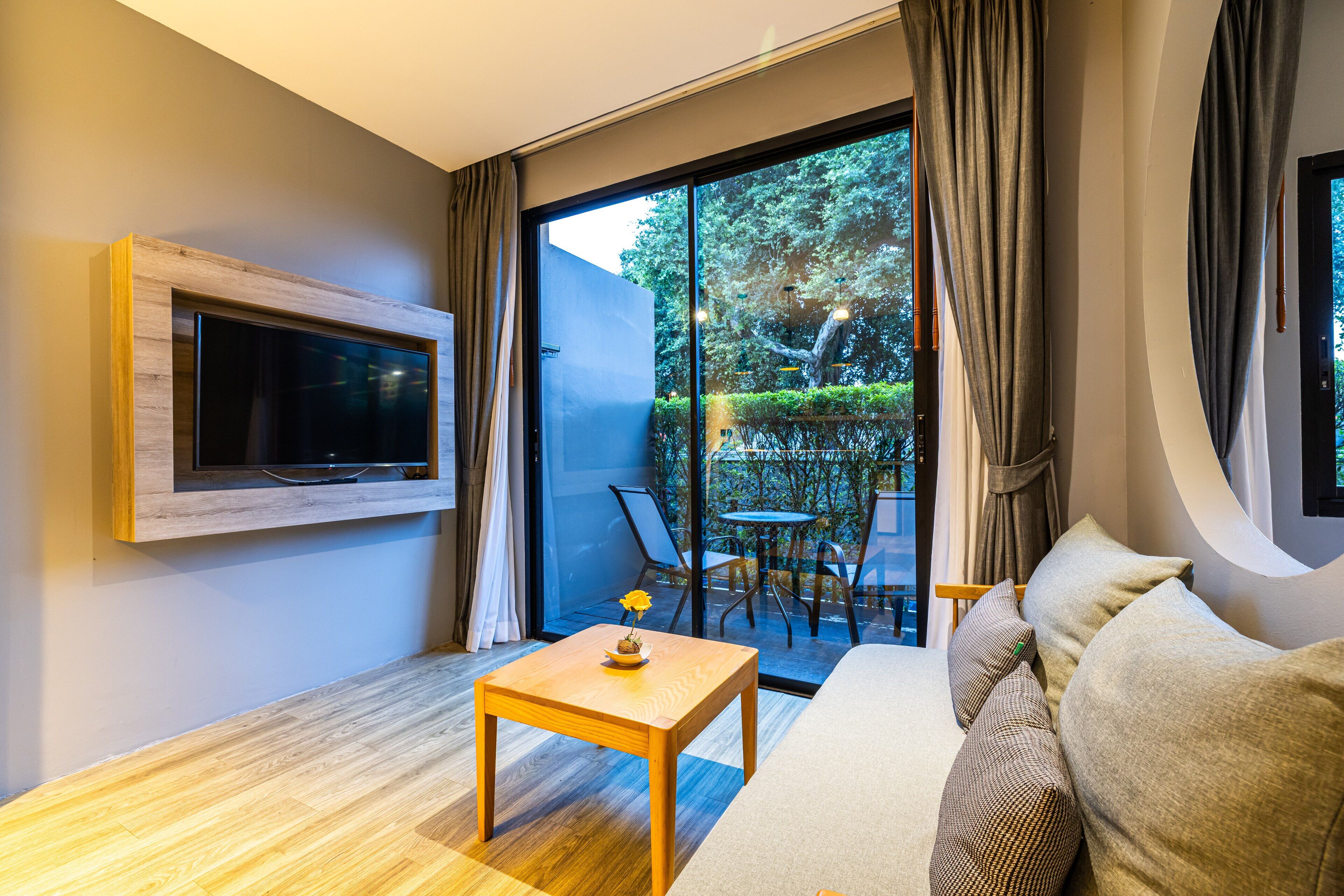 A modern living room with a flat-screen TV mounted on the wall, a wooden coffee table with a flower vase, a sofa with patterned cushions, and a sliding glass door leading to an outdoor seating area with chairs and a table.