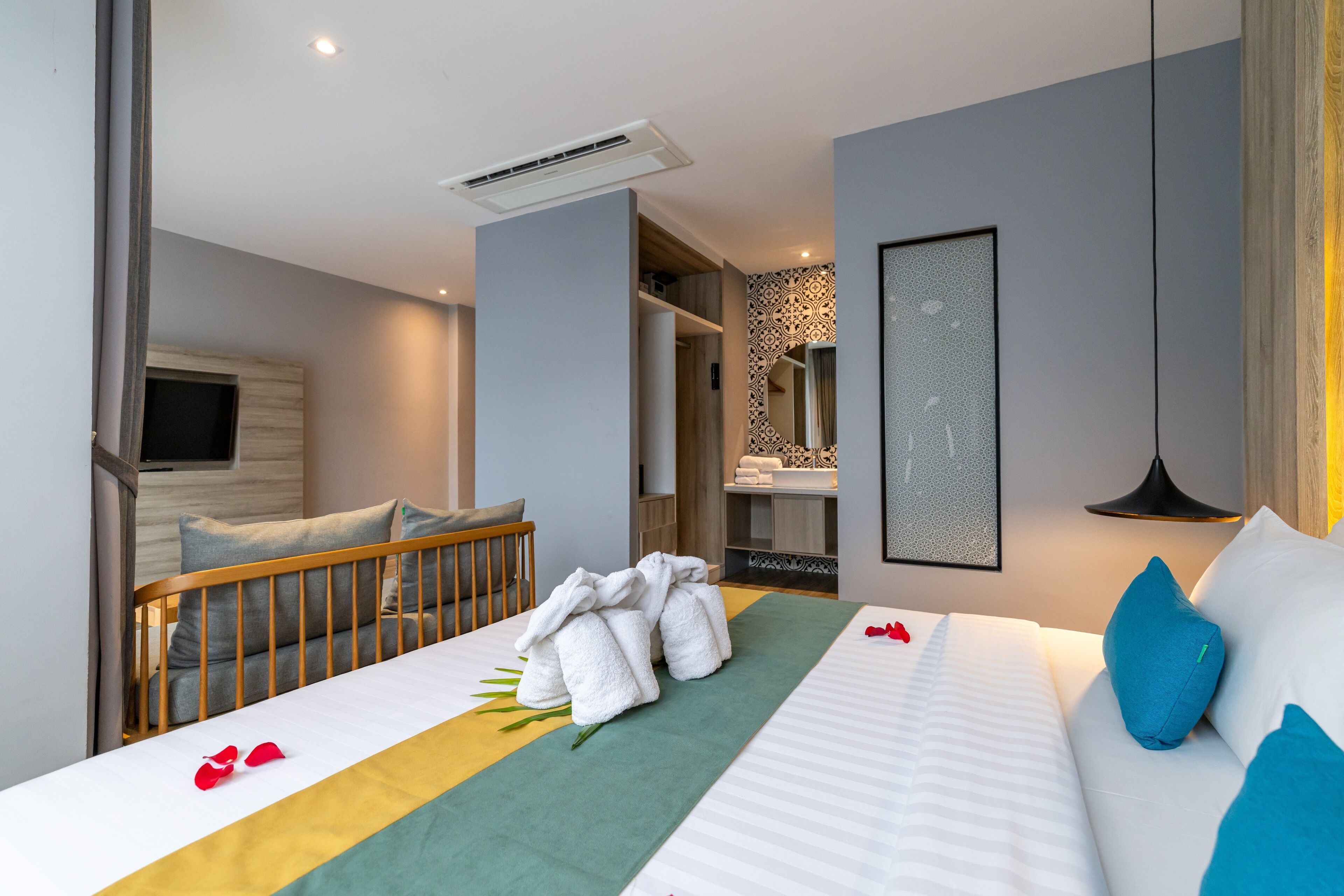 A modern hotel room with a neatly made bed, a wooden crib, a bathroom with a mirror, and a hallway leading to another room.