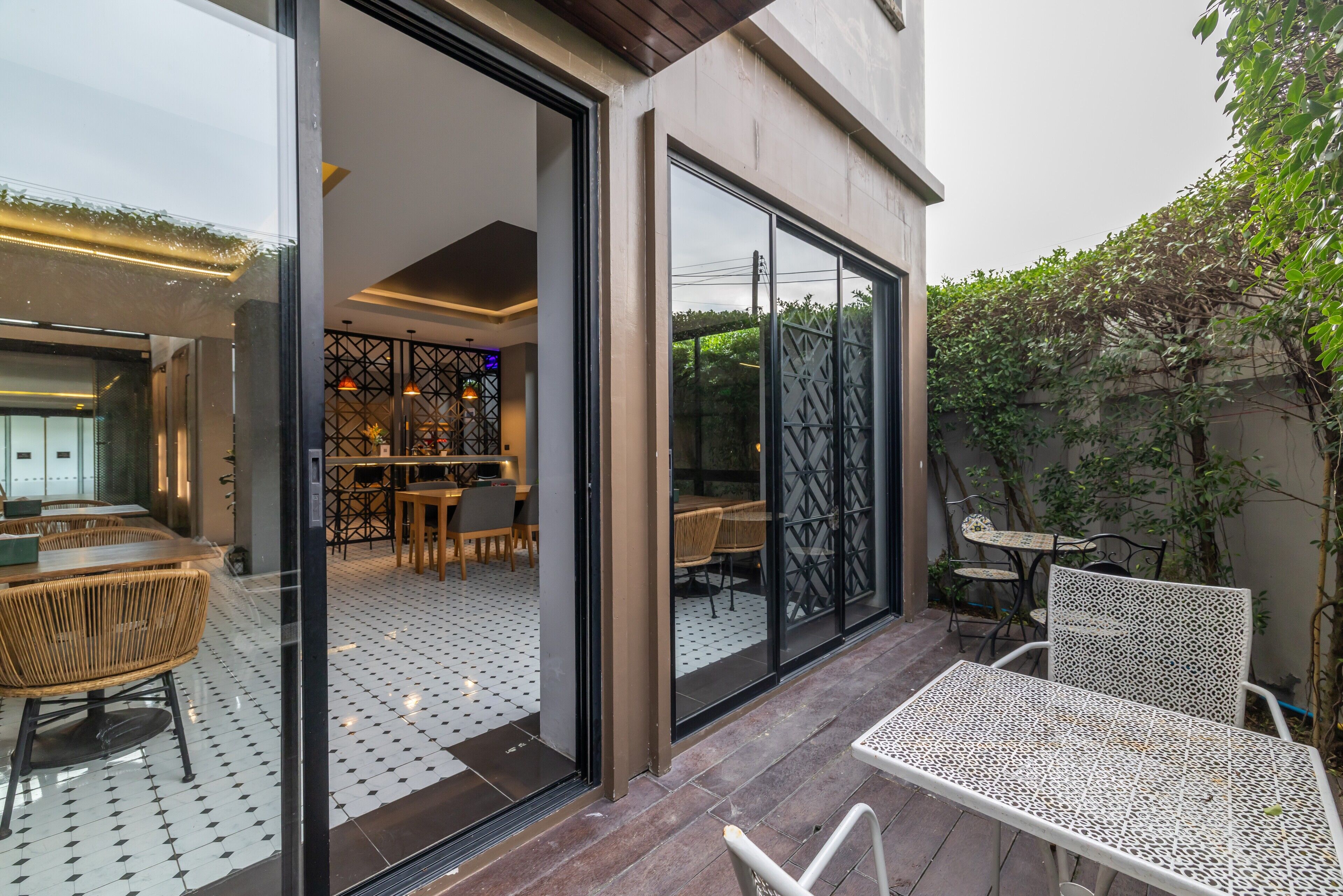 A modern outdoor patio with a dining area, a kitchen visible through glass doors, and a garden area with chairs and tables.