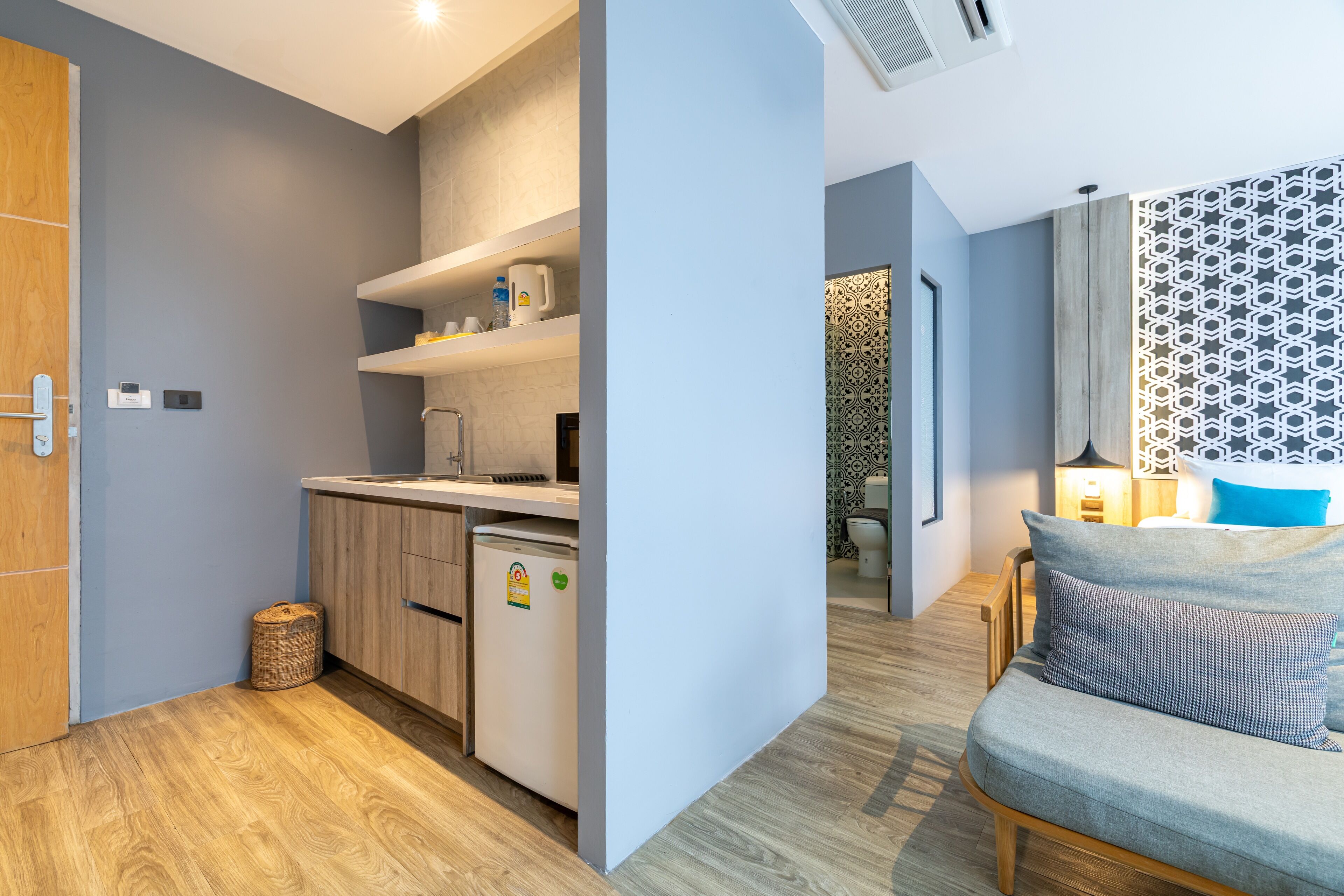 Cocina compacta con refrigerador, microondas y gabinetes de madera. Hay una cesta de mimbre en el suelo y una pequeña mesa con una lámpara.