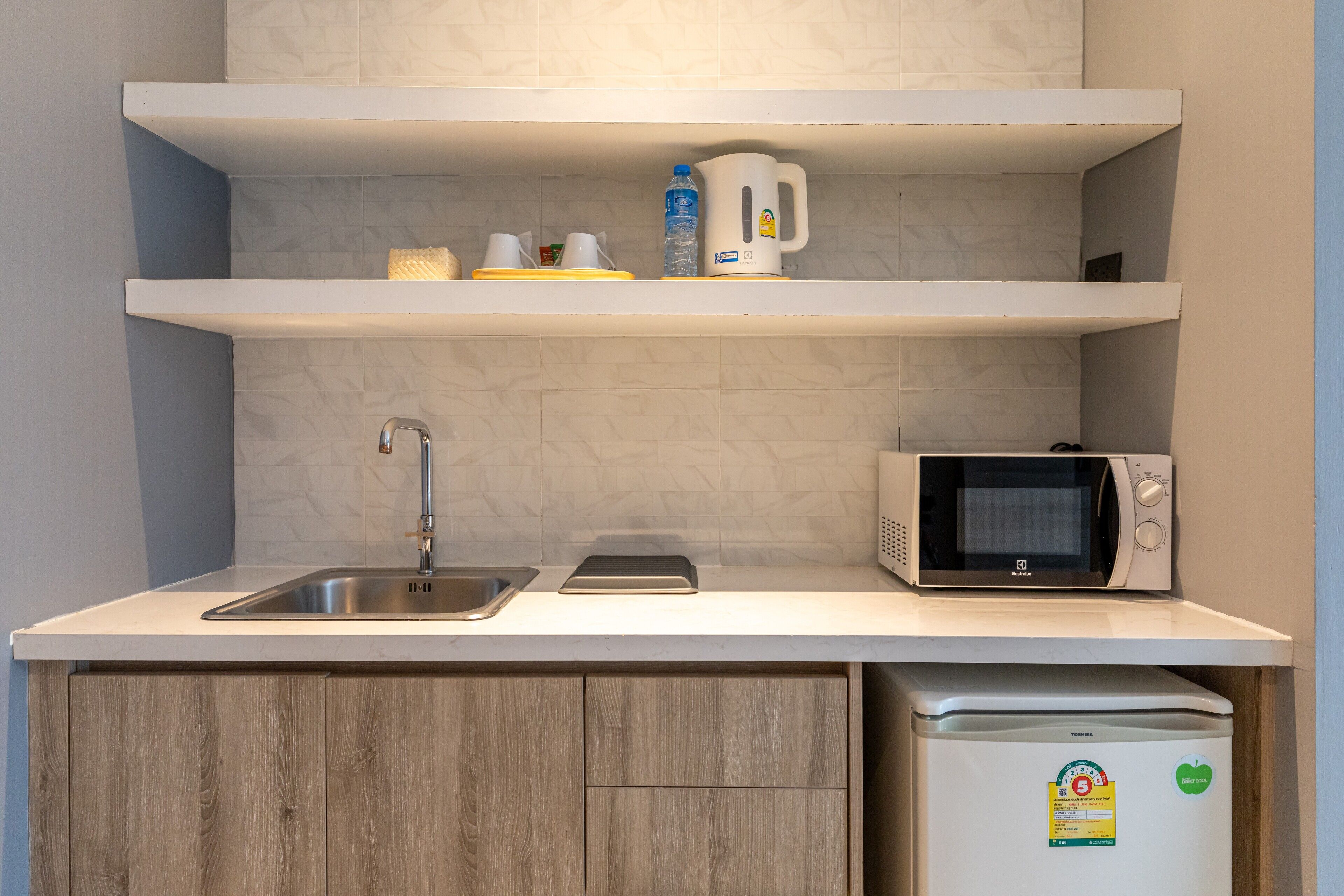 Une cuisine compacte équipée d’un évier, d’un four à micro-ondes et d’un réfrigérateur, avec des plans de travail blancs et des meubles de rangement en bois.