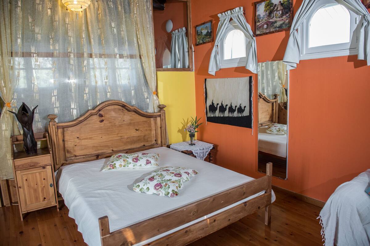Uma cama de madeira com almofadas florais, uma mesa de cabeceira com um vaso e um espelho na parede.