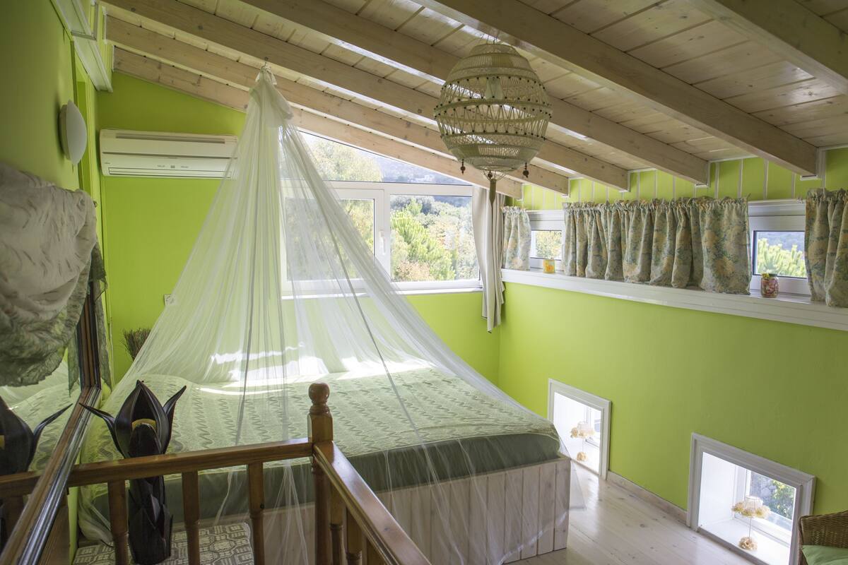 Quarto com cama sob um dossel, escada de madeira e vista para o verde lá fora.