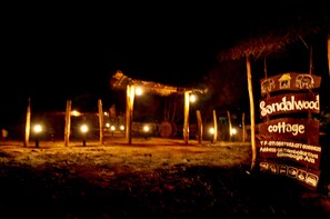 Escena nocturna de una estructura de madera rústica que dice 'Sandalwood Cottage', con un número de contacto y una dirección.