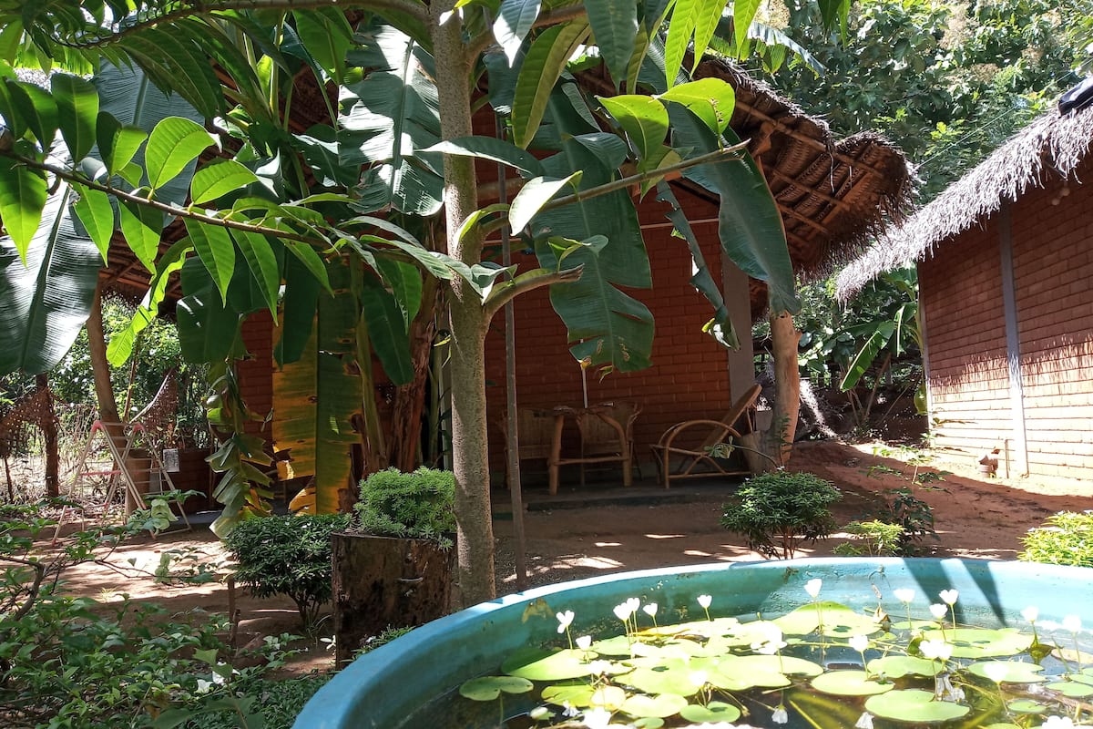 Eine rustikale Hütte mit strohgedecktem Dach, umgeben von üppigem Grün und einem kleinen Teich mit Seerosen.
