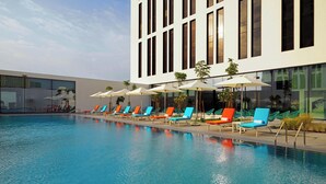 A swimming pool with colorful lounge chairs and umbrellas, surrounded by a modern building with large windows. - Aloft by Marriott Me’aisam, Dubai (Dubai)