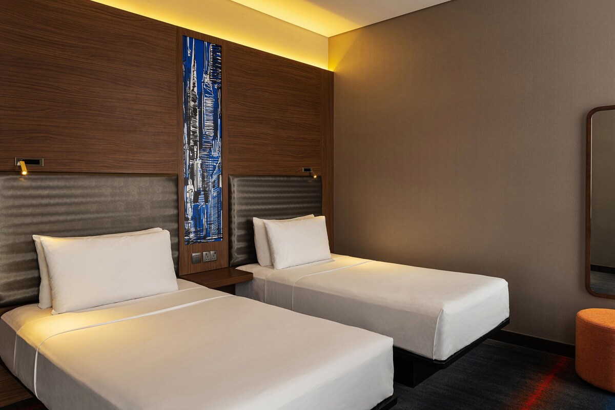 A hotel room with two single beds, a large window with a cityscape view, a wooden headboard, and a small round table.