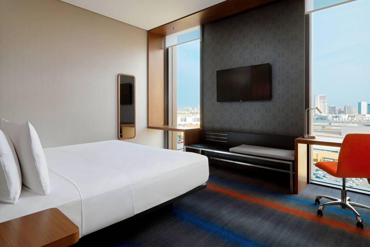 A hotel room with a large bed, a desk with a chair, a flat-screen TV mounted on the wall, and a large window offering a city view.