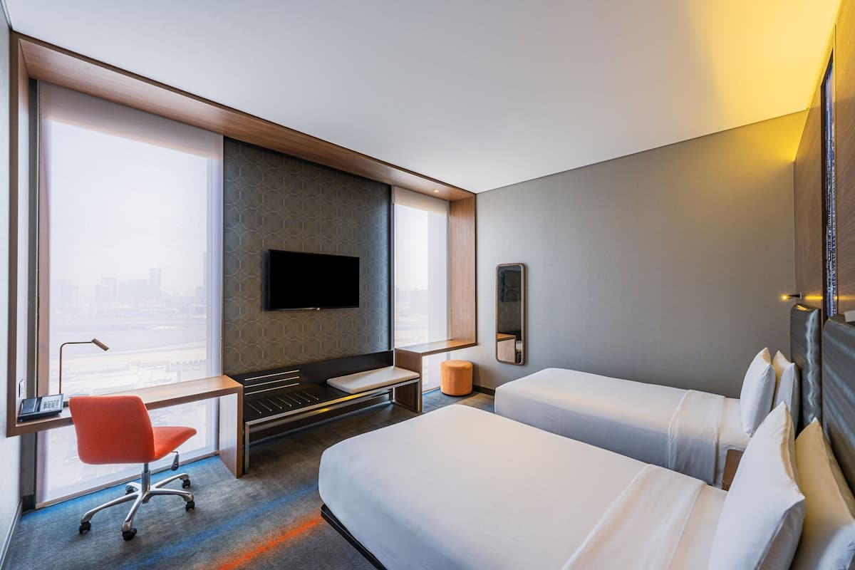 A modern hotel room with a large window, a desk with a chair, a flat-screen TV, a bed, and a bedside table.