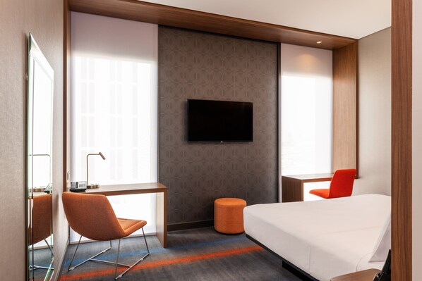 A hotel room with a bed, a television mounted on the wall, a desk with a chair, and a window with blinds.