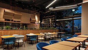 A modern restaurant interior with wooden tables and blue chairs, a bar area with shelves of bottles, and a view through large windows. - Aloft by Marriott Me’aisam, Dubai (Dubai)