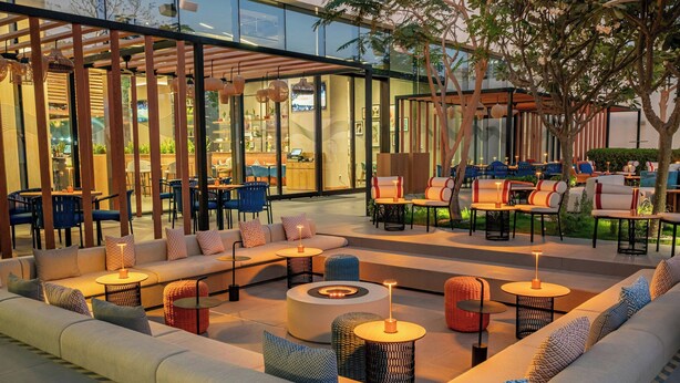 An outdoor seating area with sofas, tables, and chairs, surrounded by glass walls and trees. - Aloft by Marriott Me’aisam, Dubai (Dubai)