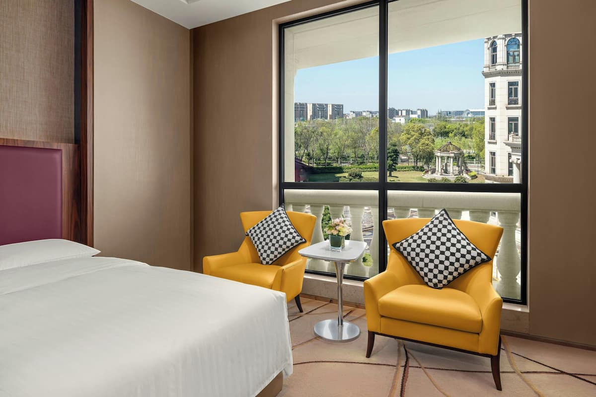 A hotel room with a bed, two yellow armchairs, a small table, and a large window offering a city view.