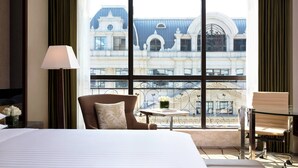A hotel room with a large bed, a chair, a small table with a vase of flowers, and a view of a historic building through large windows.