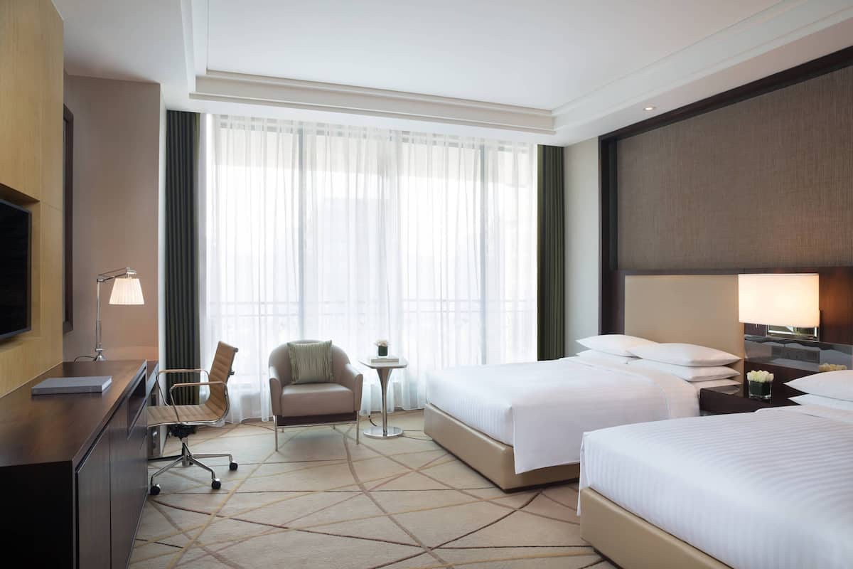 A hotel room with two beds, a desk with a computer, a chair, a small table with a lamp, and a large window with sheer curtains.
