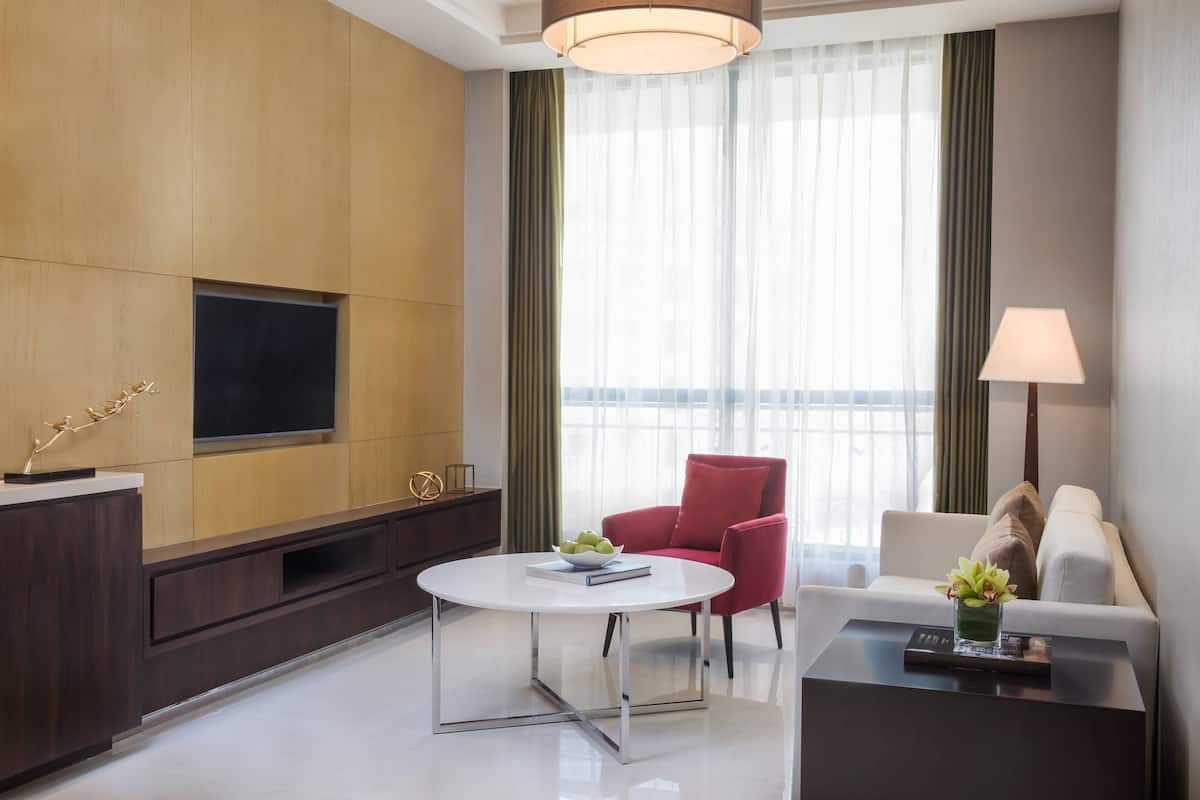 A modern hotel room with a flat-screen TV mounted on a wooden panel wall, a round white coffee table, a red armchair, a white sofa, and a small table with a lamp and a vase of flowers.